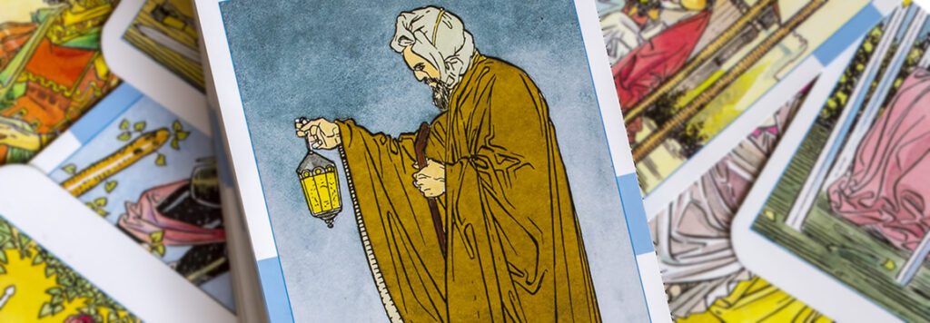 A tarot card featuring a hooded figure holding a lantern, representing the Hermit. The card is placed among a spread of other colorful tarot cards, each partially visible and displaying intricate designs.