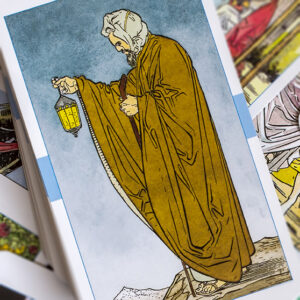 A tarot card depicting an elderly man with a long beard wearing a hooded robe. He holds a staff in one hand and a lit lantern in the other, standing on rocky terrain. The background is a pale blue sky.