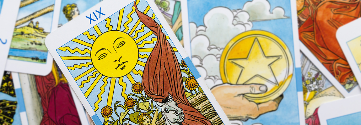 Close-up of a spread of tarot cards, with the Sun card prominently featured in the center. The card depicts a bright yellow sun with a face, radiating light, surrounded by sunflowers. Other cards with various symbols are partially visible in the background.