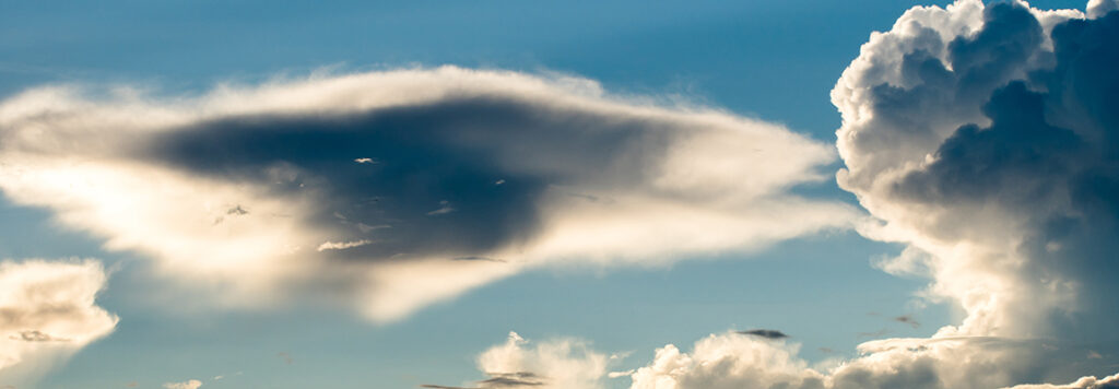 A vast sky with a dramatic cloudscape features a large, dark, UFO-shaped cloud on the left, contrasting with bright, fluffy cumulus clouds on the right, all set against a blue sky.