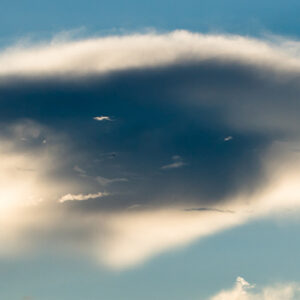 A fluffy, dark gray cloud with a soft, rounded shape stretches across a blue sky. Sunlight subtly highlights the cloud's edges, creating a dramatic contrast between the light and dark areas.