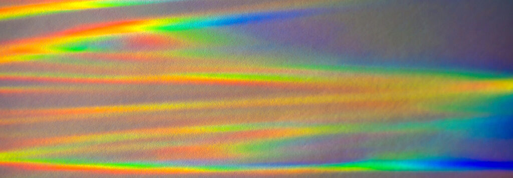 Colorful rainbow light patterns are projected onto a textured surface, creating a vibrant display of overlapping lines and gradients. The spectrum includes vivid reds, oranges, yellows, greens, blues, and purples.