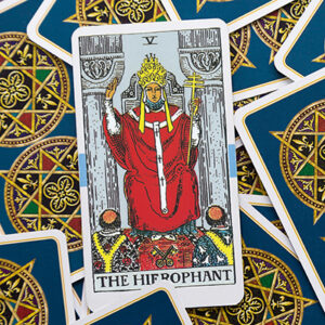 A tarot card called "The Hierophant" is placed on a pile of cards with intricate, colorful designs. The card depicts a figure in red robes and a crown, holding a cross, symbolizing spiritual guidance and tradition.