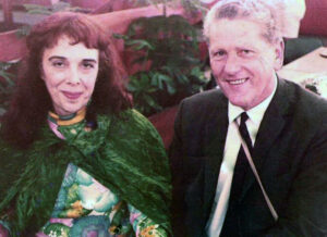 Reverend Ann Davies  in a green floral dress and Reverend Will Chesterman in a suit sit side by side, smiling, in a warmly lit room with plants in the background.