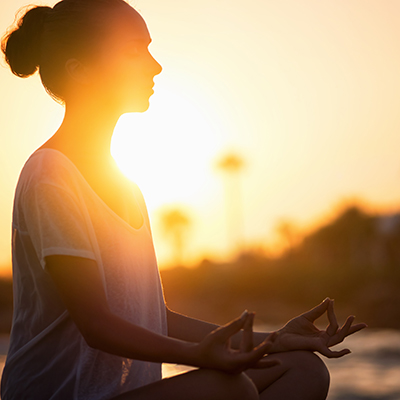 Silhouette of a person meditating outdoors at sunset, sitting cross-legged with hands in a mudra gesture, with the sun shining brightly behind them.