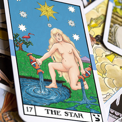 A close-up of the tarot card "The Star," depicting a nude woman kneeling by water, pouring liquid from two jugs. Bright stars fill the blue sky, and green fields are in the background. Other tarot cards are partially visible beneath.