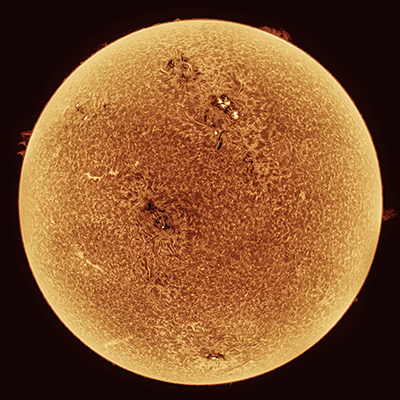 Close-up image of the Sun, showing its textured surface with several dark sunspots and bright regions, surrounded by a glowing, orange aura against a dark background.