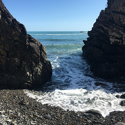 Waves wash onto a rocky beach framed by two large, dark cliffs under a clear blue sky. The ocean stretches out to the horizon in the background.