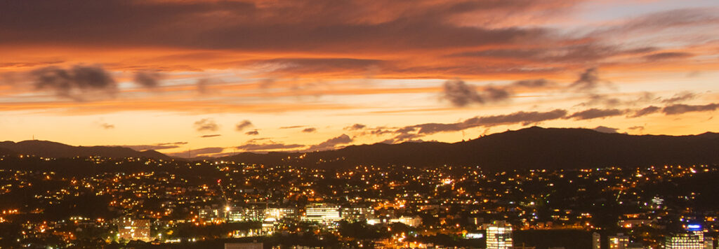 Wellington Harbour and City: A cityscape at dusk with twinkling lights and dark silhouettes of hills under a dramatic orange and purple sunset sky filled with scattered clouds.