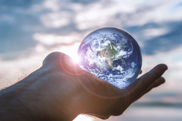 A close-up of a hand holding a small, realistic model of Earth with the sun shining in the background and a cloudy sky.