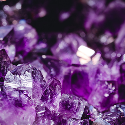Close-up of purple amethyst crystals, showing sharp, angular facets and a vibrant, translucent texture with highlights and reflections.
