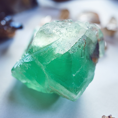 A close-up of a rough green crystal with translucent edges, resting on a white surface. The crystal has uneven, jagged angles and appears to catch the light, highlighting its texture and color variations.