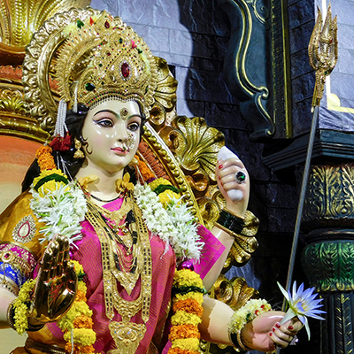 A colorful statue of a Hindu goddess adorned with an ornate crown, elaborate jewelry, and floral garlands, holding a trident and a lotus flower, set against a decorative background.