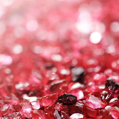 A close-up view of numerous small, rough red gemstones or crystals, with a sparkling and slightly blurred background that creates a vibrant, shimmering effect.