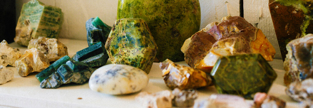Colorful, variously shaped crystals and minerals are displayed on a white shelf. The collection includes green, yellow, and multicolored stones, some rough and some with smooth, polished surfaces.
