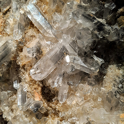 Close-up of clear quartz crystals clustered together on a rocky, beige-brown surface, showing sharp, angular shapes and transparent, reflective edges.